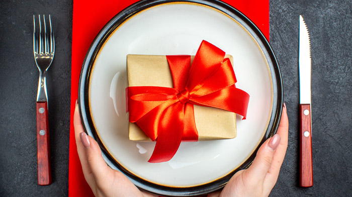 リボンのかかった小さなギフトボックスの置かれたランチョンマットの上の皿に両手を添えた、グルメギフトのイメージ画像